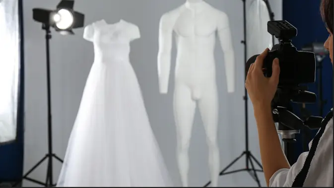 How to Ghost Mannequin Photography A photographer, seen from the side, holds a DSLR camera to their eye, focusing on two white mannequins in a photo studio